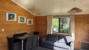 Um quarto acolhedor com paredes de madeira, um sofá cinzento, uma mesa de jantar com cadeiras e uma janela grande com cortinas.