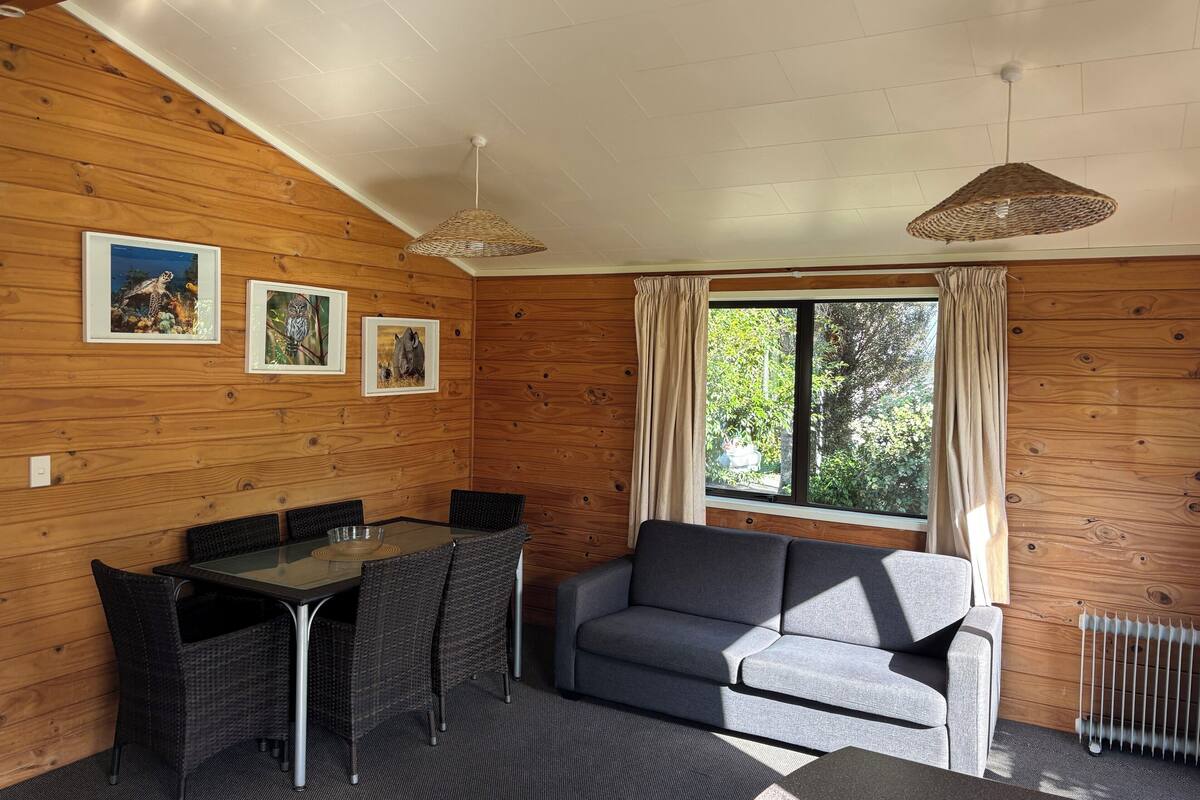 A cozy room with wooden walls, a grey sofa, a dining table with chairs, and a large window with curtains.