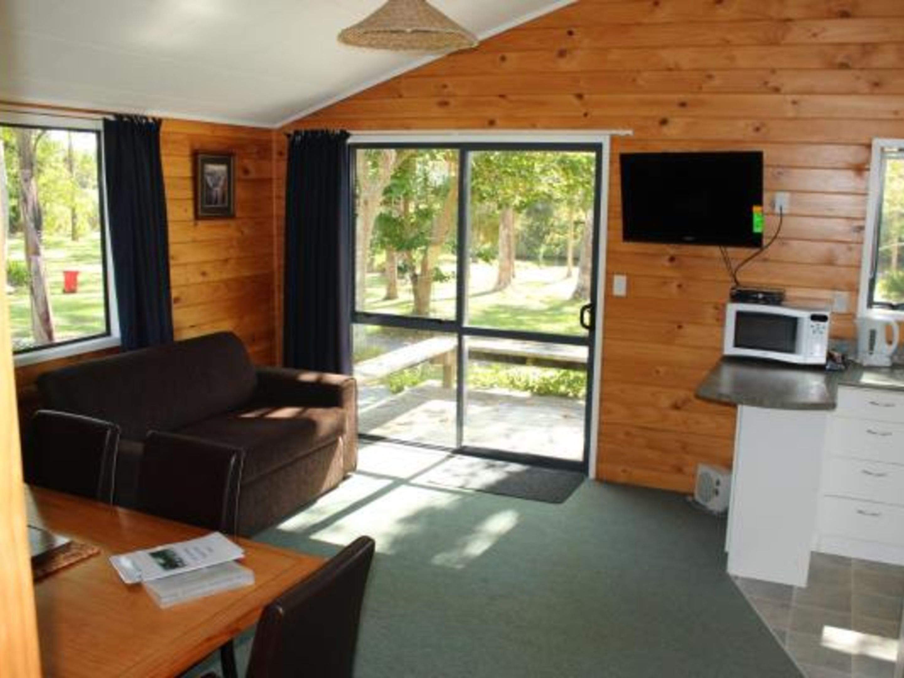 Eine Hütte mit Holzvertäfelung, einem Sofa, einem Tisch mit Papieren, einem an der Wand befestigten Fernseher und einer Küchenzeile mit einer Mikrowelle.