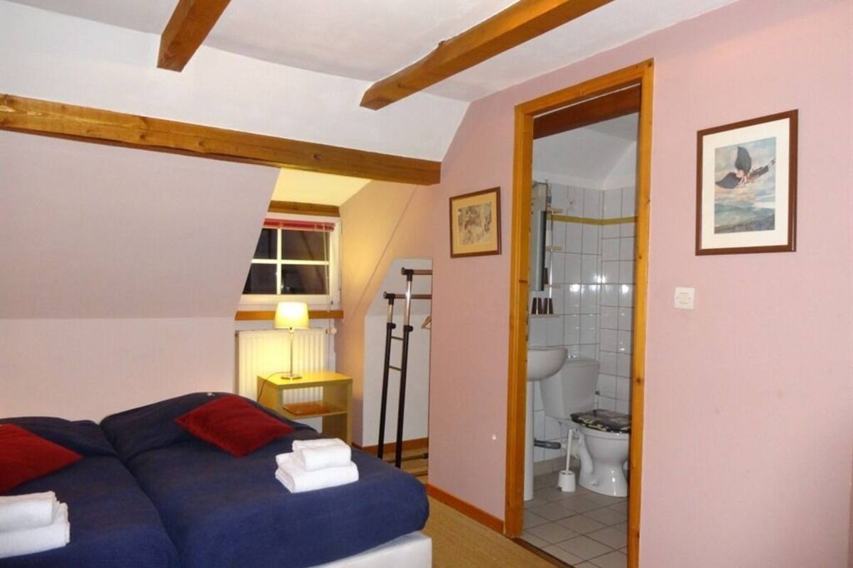 A bedroom with two beds, a wooden ceiling, a bathroom visible through an open door, and a small shelf with a lamp.