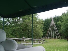 Un área de estar techada con vista a un bosque y una estructura de madera al fondo.