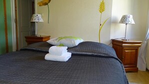 A neatly made bed with a dark quilt, two bedside lamps, and a wall with a wheat stalk and flower decoration. - Pension Linstant (Le Hohwald)