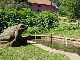 Una estatua de rana de piedra en un estanque, con una casa y follaje al fondo.