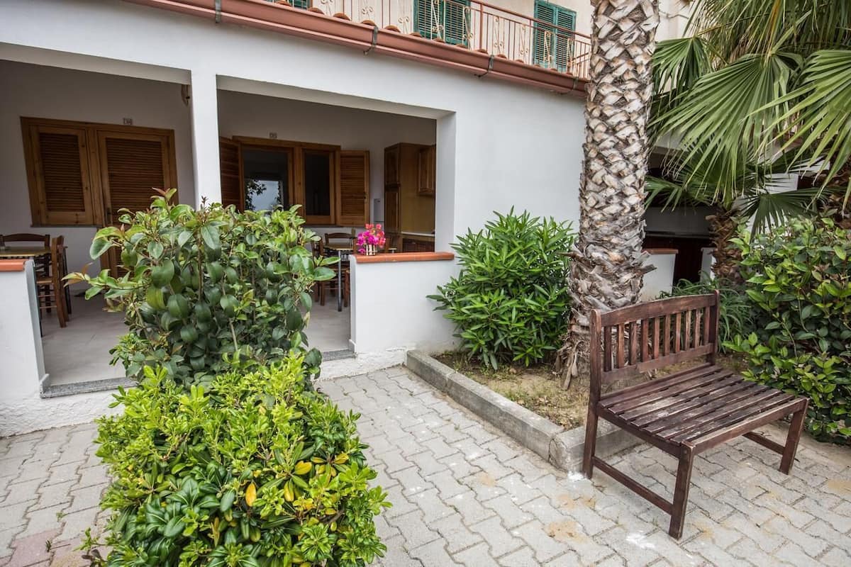 A courtyard with a wooden bench, potted plants, and a white building with wooden shutters.