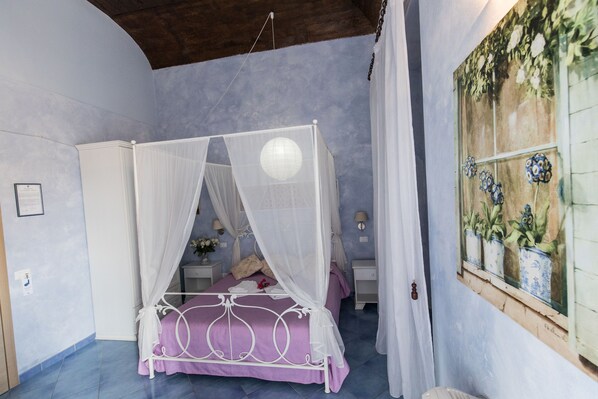 A bedroom with a four-poster bed, a nightstand, a wall-mounted lamp, and a window with a floral view. - Blu Tropea Maison B&B (Tropea)