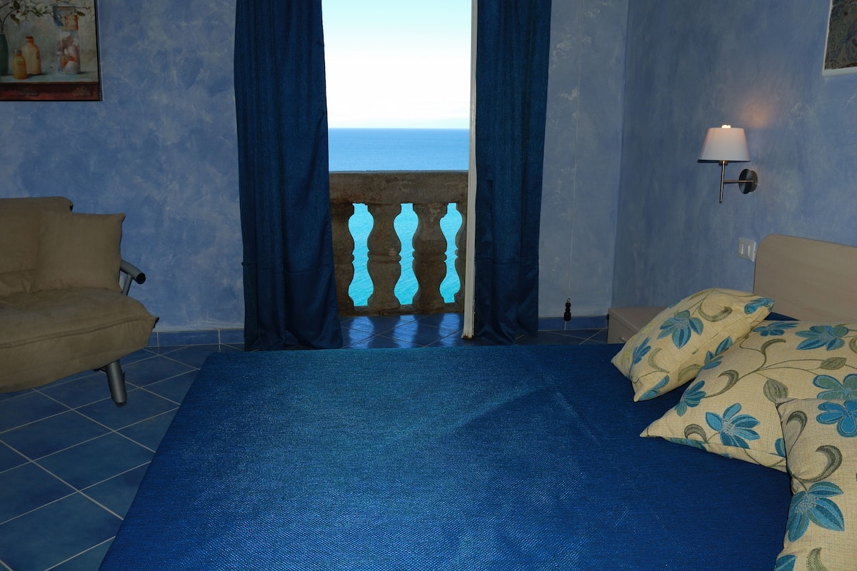 A bedroom with a large bed, a sofa, and a view of the sea through a window with curtains.