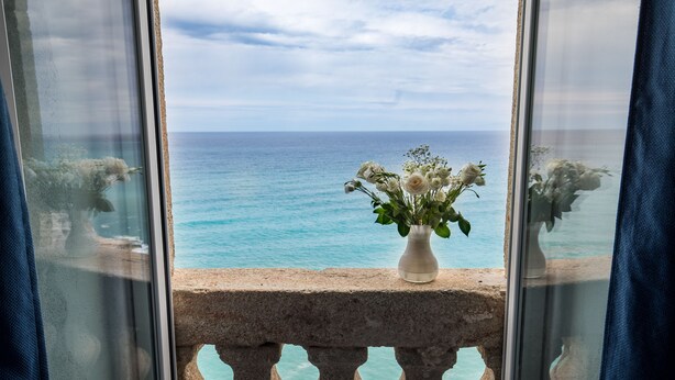 A balcony with a stone balustrade overlooking a turquoise sea. A white vase with flowers is placed on the balustrade.