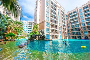 A swimming pool surrounded by multi-story buildings with balconies, palm trees, and a rocky feature in the water. - Atlantis Condo (Pattaya)