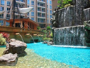 A swimming pool with a waterfall feature, surrounded by rocks and palm trees, with a building in the background. - Atlantis Condo (Pattaya)