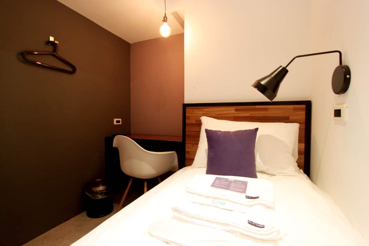 Una habitación de hotel con una cama bien hecha, una lámpara de noche, un perchero y una silla blanca.