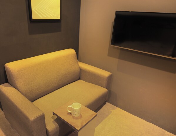 A room with a beige sofa, a small wooden table with a white mug, and a flat-screen TV mounted on the wall. - Daan Park Hotel - Hostel (Taipei)
