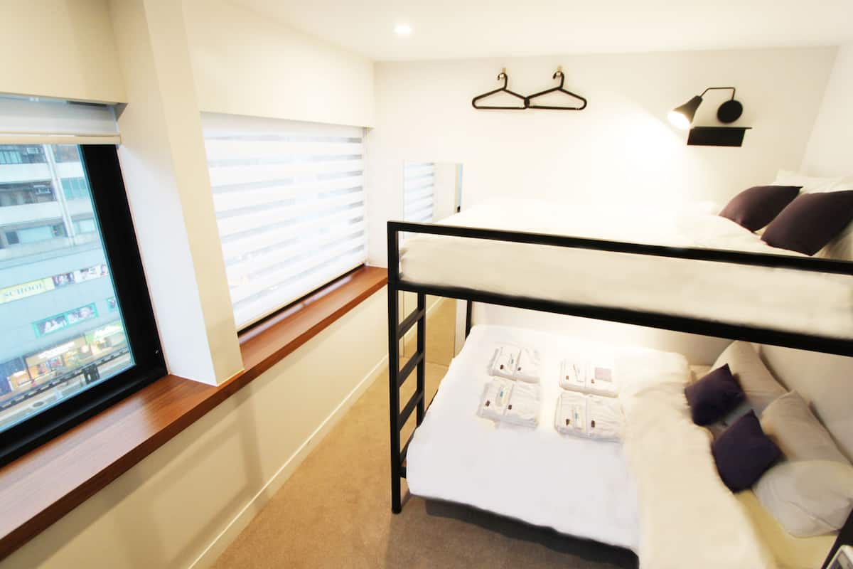 Una cama litera con colchón blanco y almohadas moradas, una ventana con persianas y una lámpara fijada a la pared.