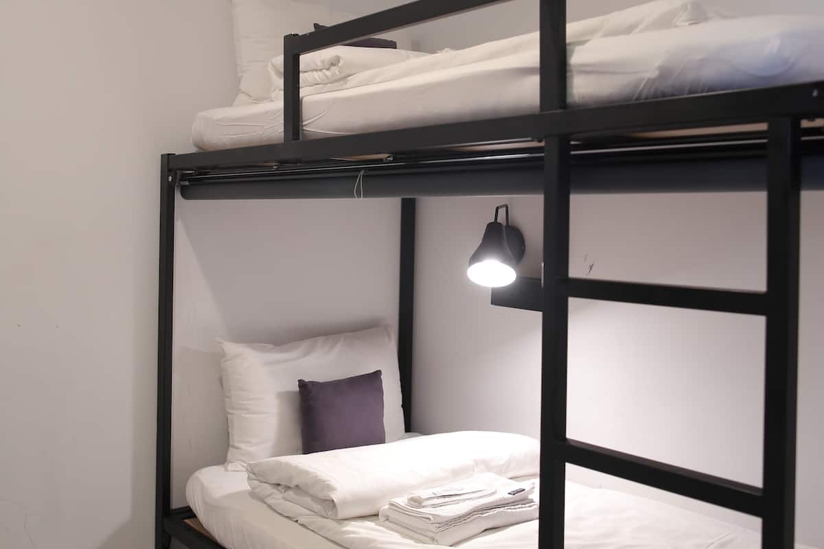Una cama individual superpuesta con estructura negra, ropa de cama blanca y una lámpara de noche.