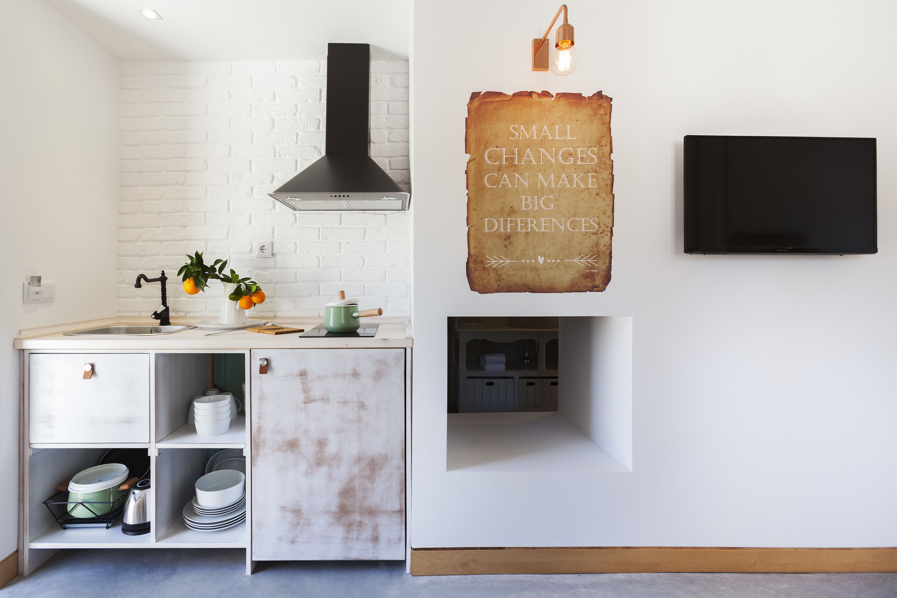 A kitchen with white brick backsplash, a black range hood, and a wall-mounted TV. There's a sign that reads "SMALL CHANGES CAN MAKE BIG DIFFERENCES" and a small shelf with dishes.