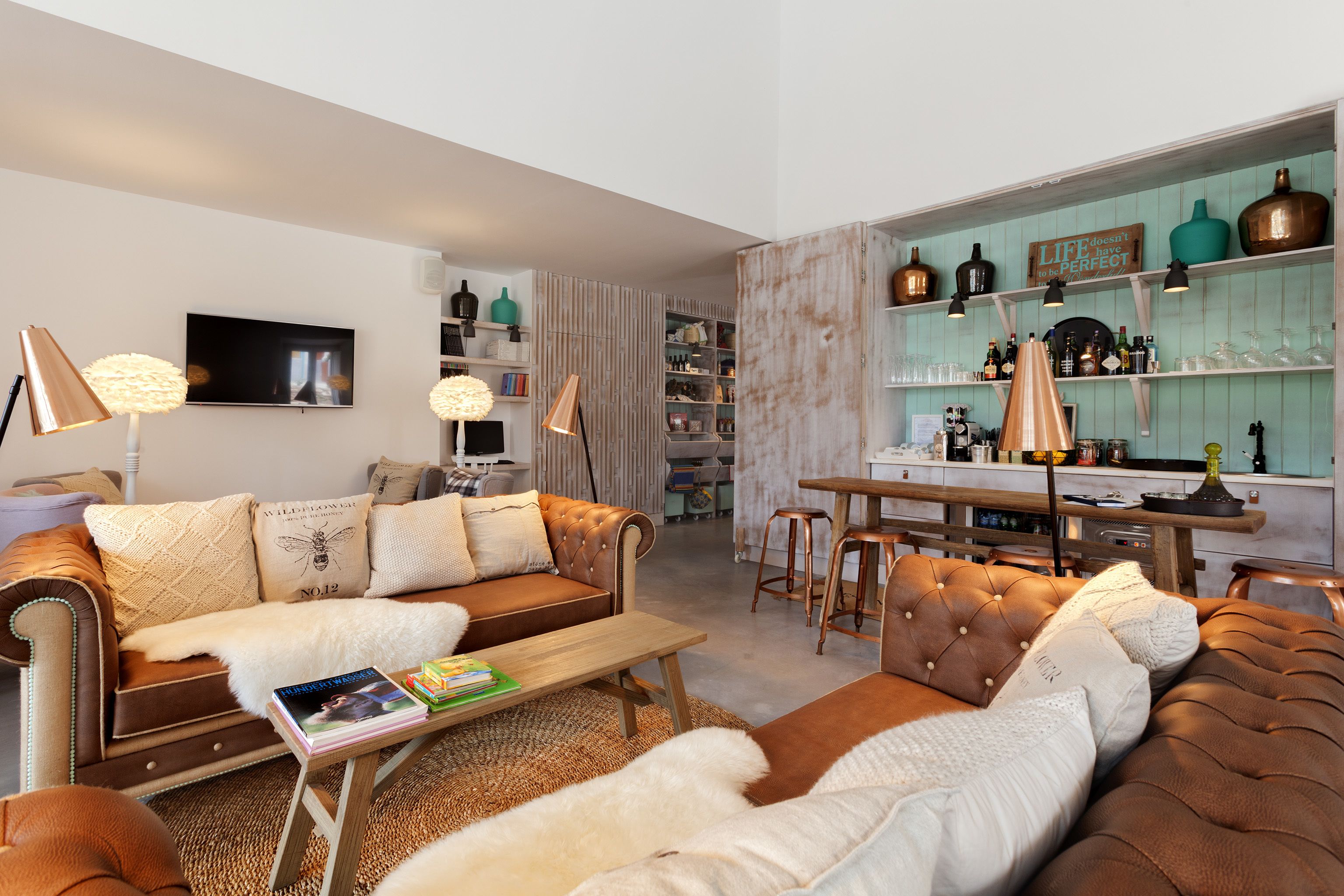 A modern living room with a brown leather sofa, a wooden coffee table, and a TV mounted on the wall. There are shelves with various items and a kitchen area in the background.
