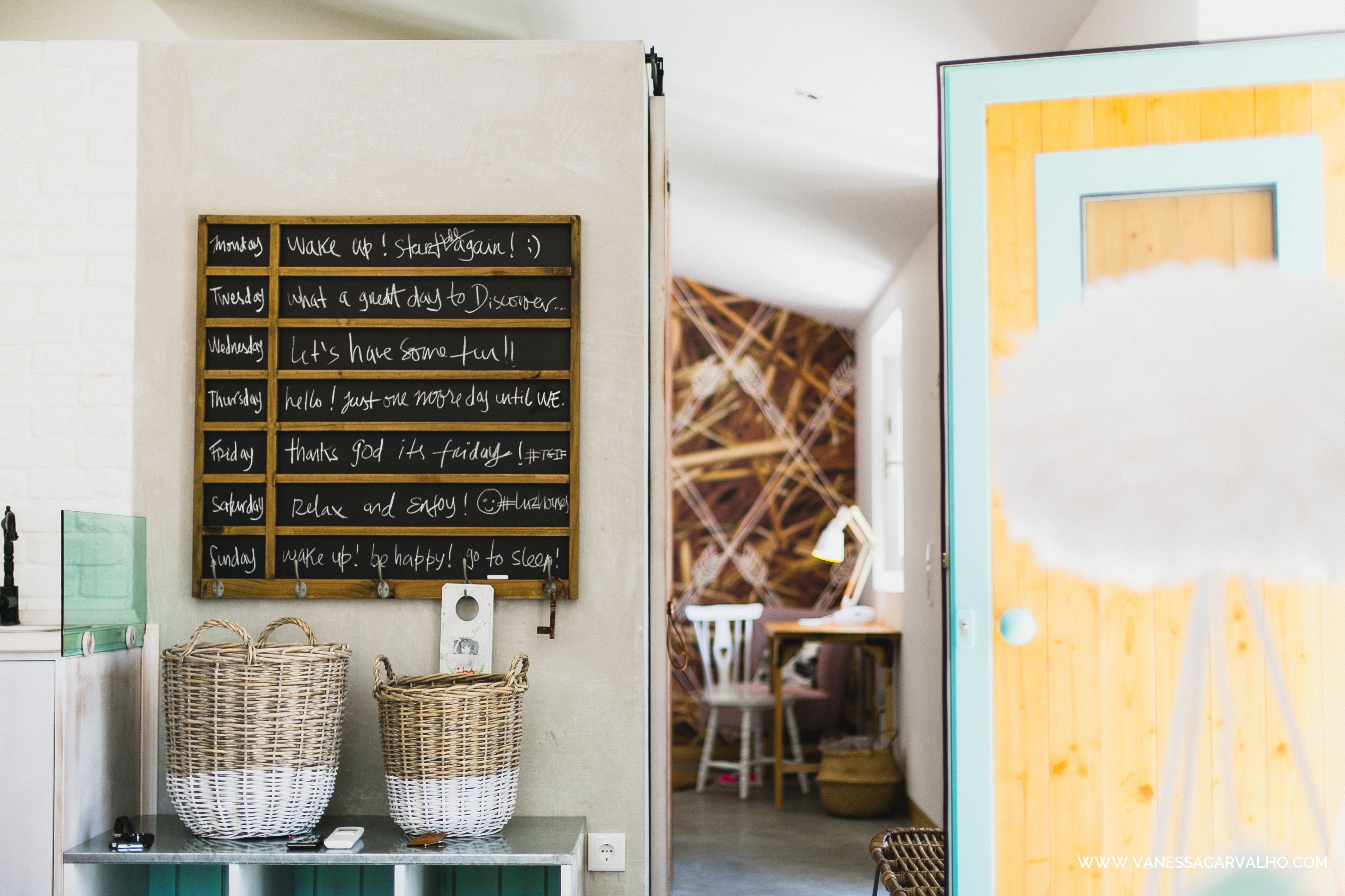 A room with a wall-mounted chalkboard displaying daily motivational messages, wicker baskets on a shelf, and a wooden door with a white fluffy object in front.