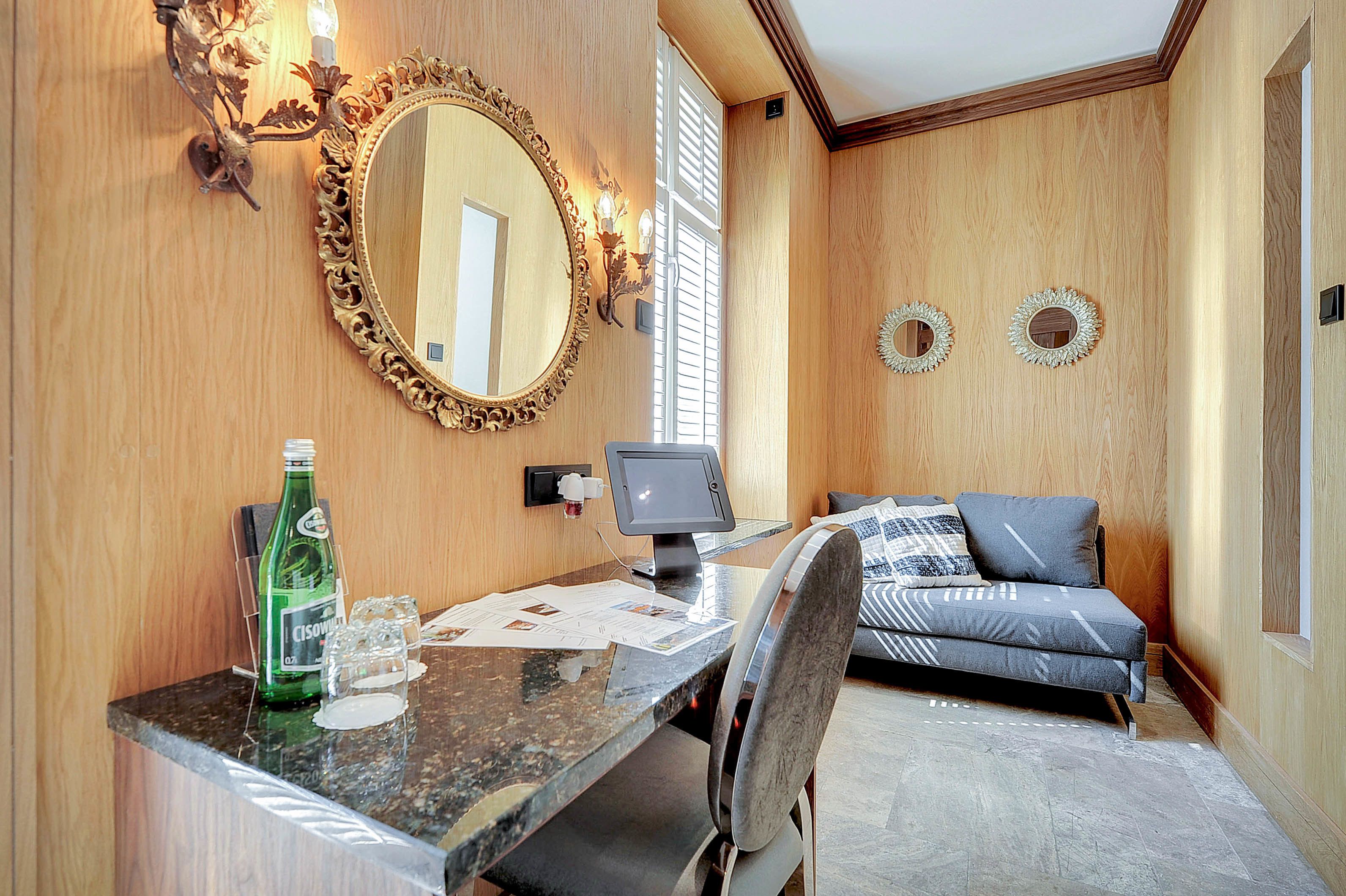 Una habitación con pared de madera, un espejo grande y ornamentado, un escritorio pequeño con una computadora, un sofá y espejos decorativos en la pared.