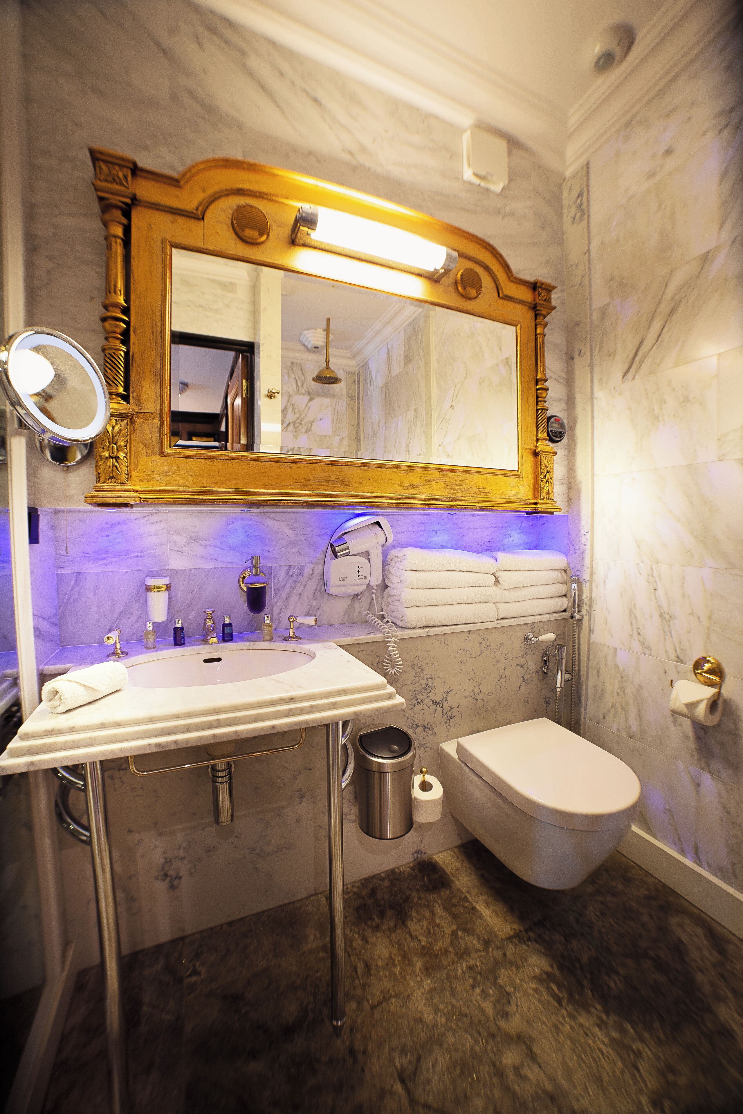 A bathroom with a marble sink, a toilet, and a shower. There is a large mirror with a gold frame above the sink.