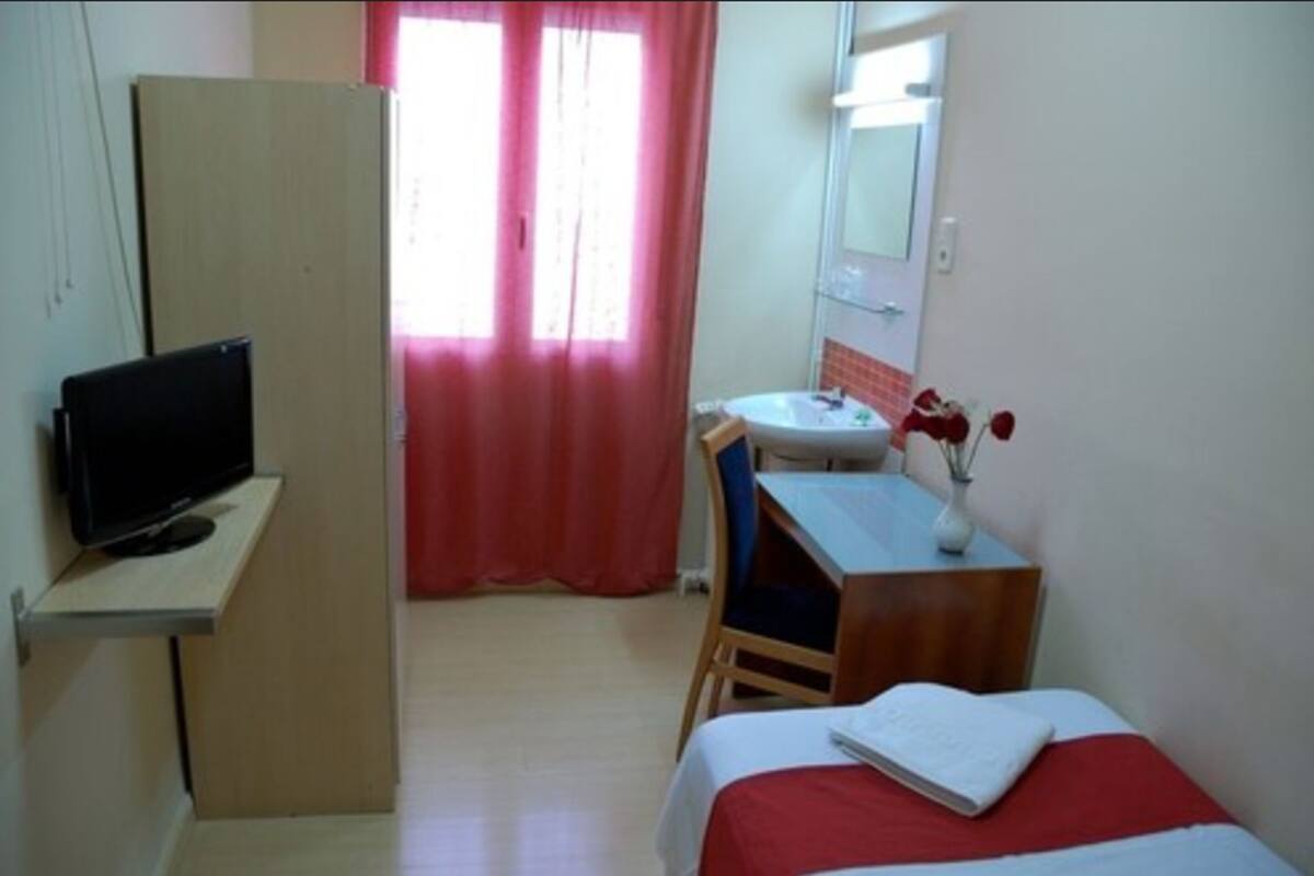A small, simple room with a bed, a desk, a chair, a TV, a sink, and a mirror.