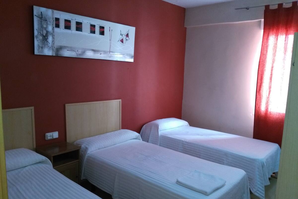 A hotel room with three single beds, a wooden headboard, a wall-mounted artwork, and a window with red curtains.