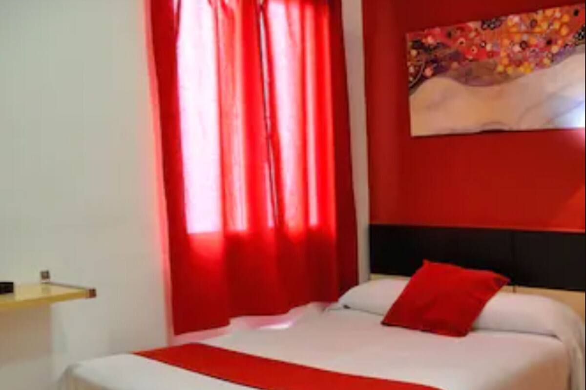 A hotel room with a bed, red curtains, a white headboard, and a painting on the wall.