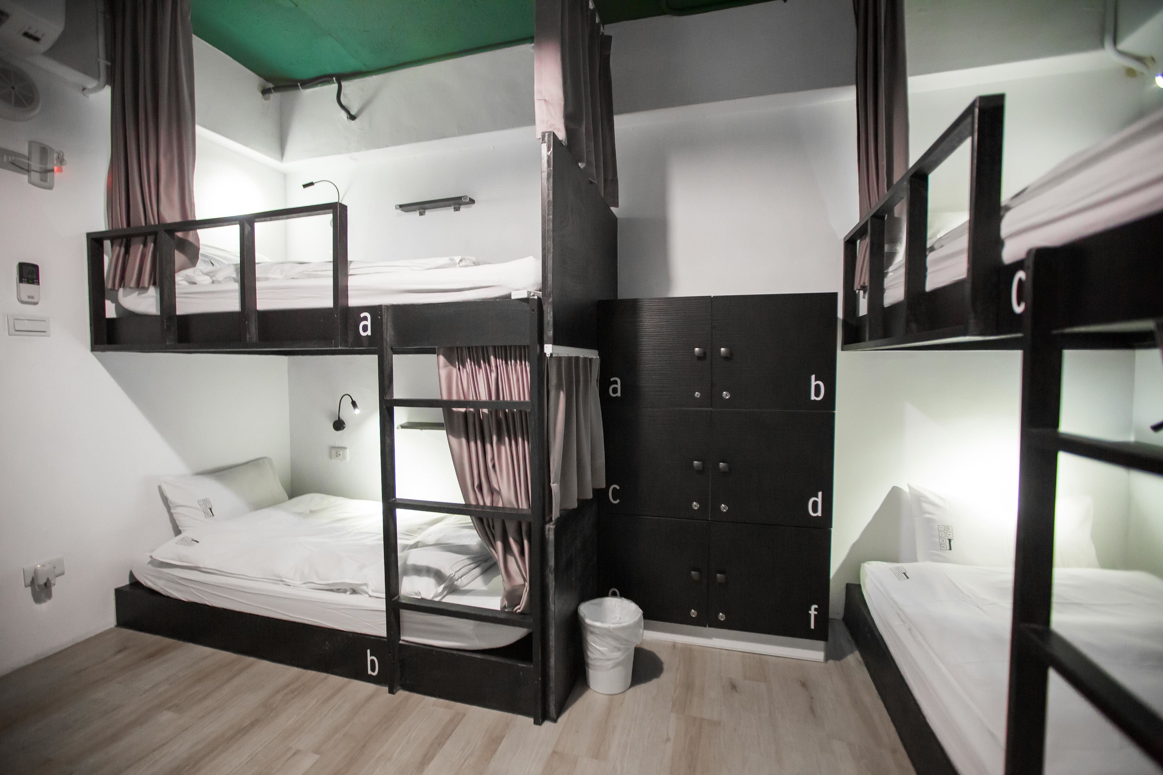 A dormitory room with bunk beds, a wardrobe, a trash bin, and a small desk with a lamp.