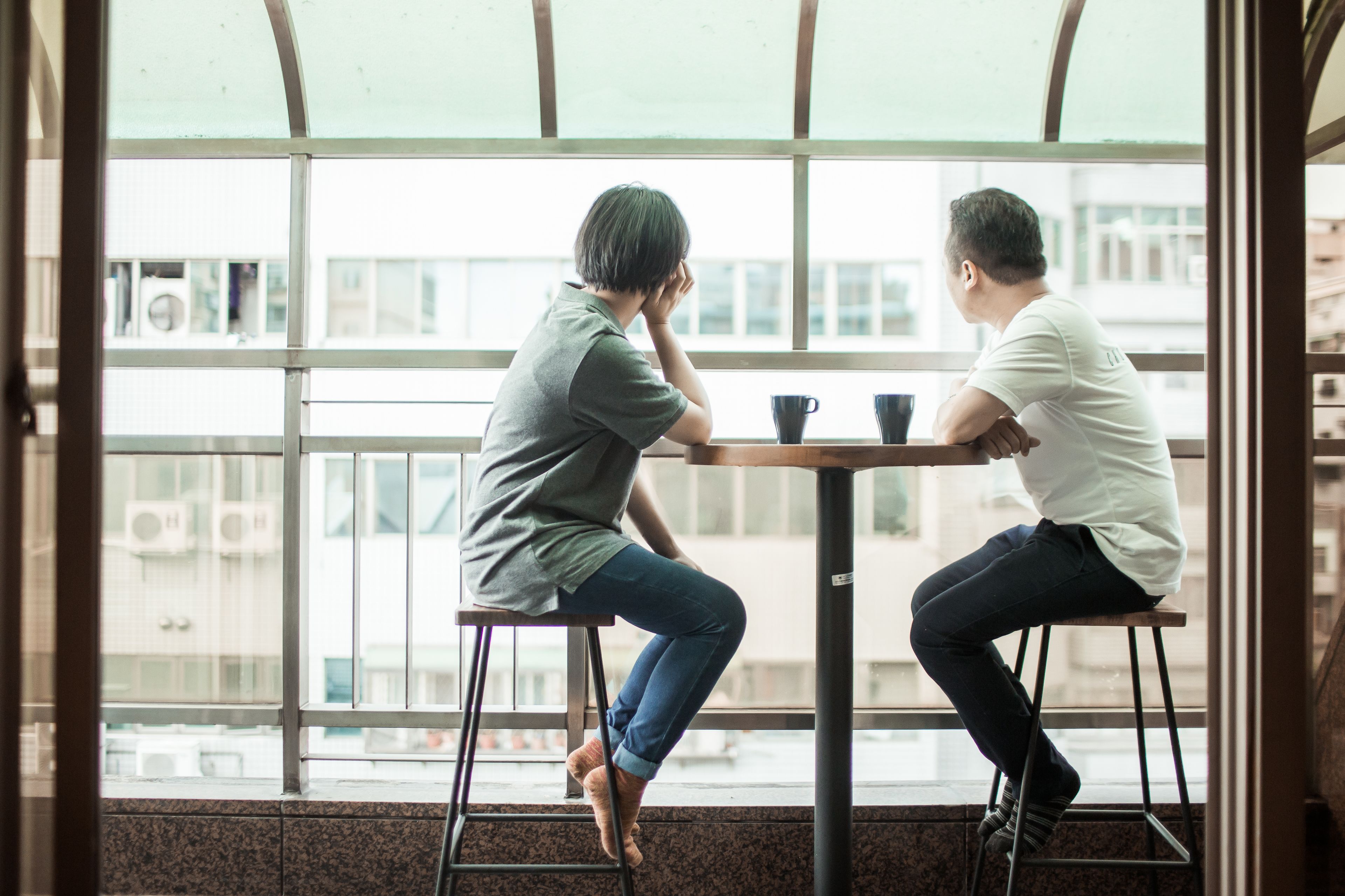 Twee personen zitten aan een tafel bij een groot raam met uitzicht op gebouwen.