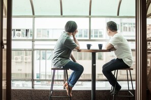 Two individuals seated at a table by a large window with a view of buildings. - We Come Hostel (Taipei)