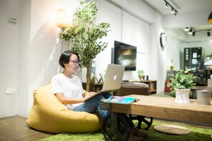A person sitting on a yellow bean bag chair using a laptop in a modern living room with a wooden table, a potted plant, and a television in the background. - We Come Hostel (Taipei)