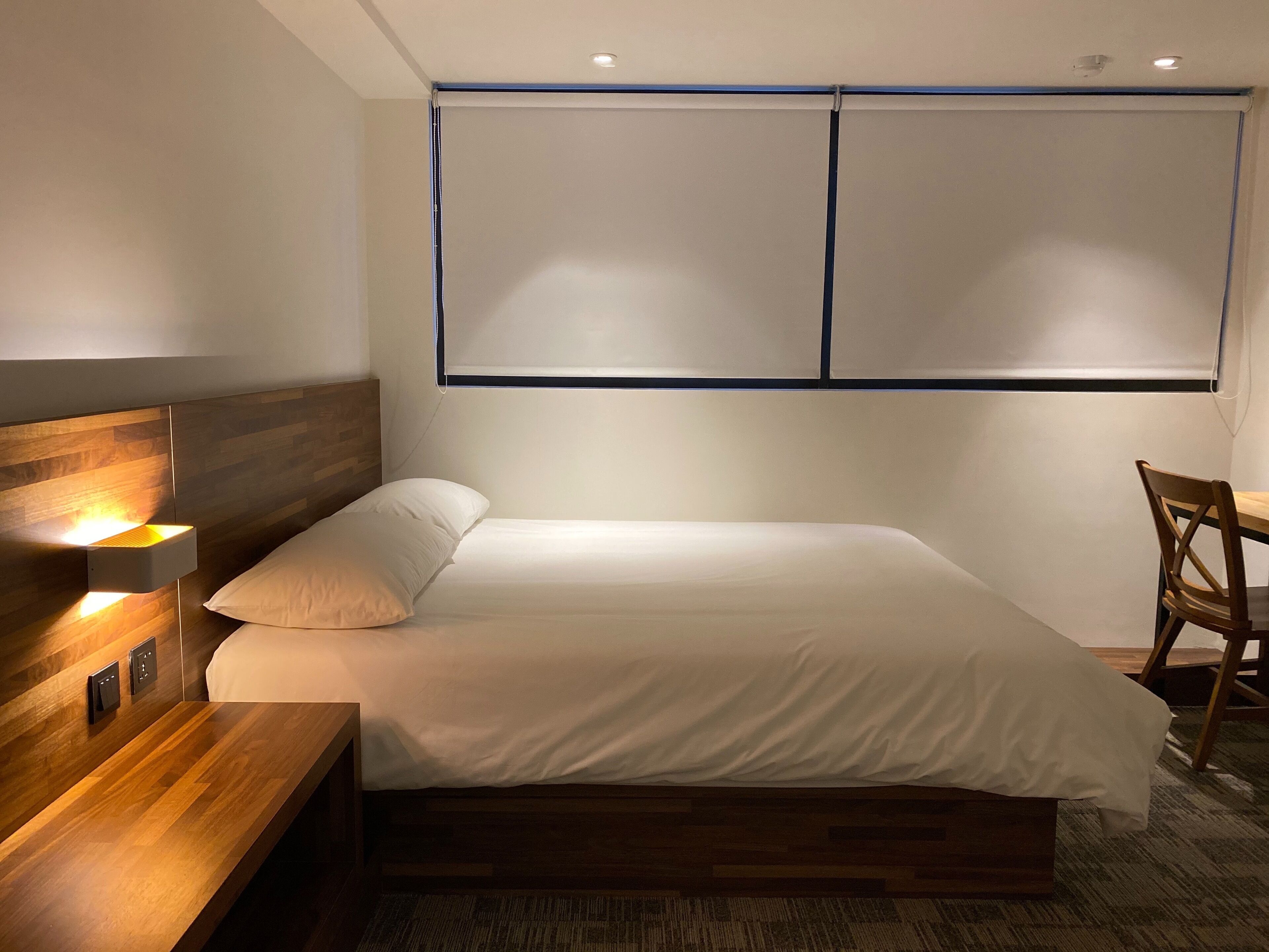 A modern bedroom with a wooden headboard, a bedside table with a lamp, a bed with white bedding, and a window with blinds.
