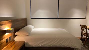 A modern bedroom with a wooden headboard, a bedside table with a lamp, a bed with white bedding, and a window with blinds.