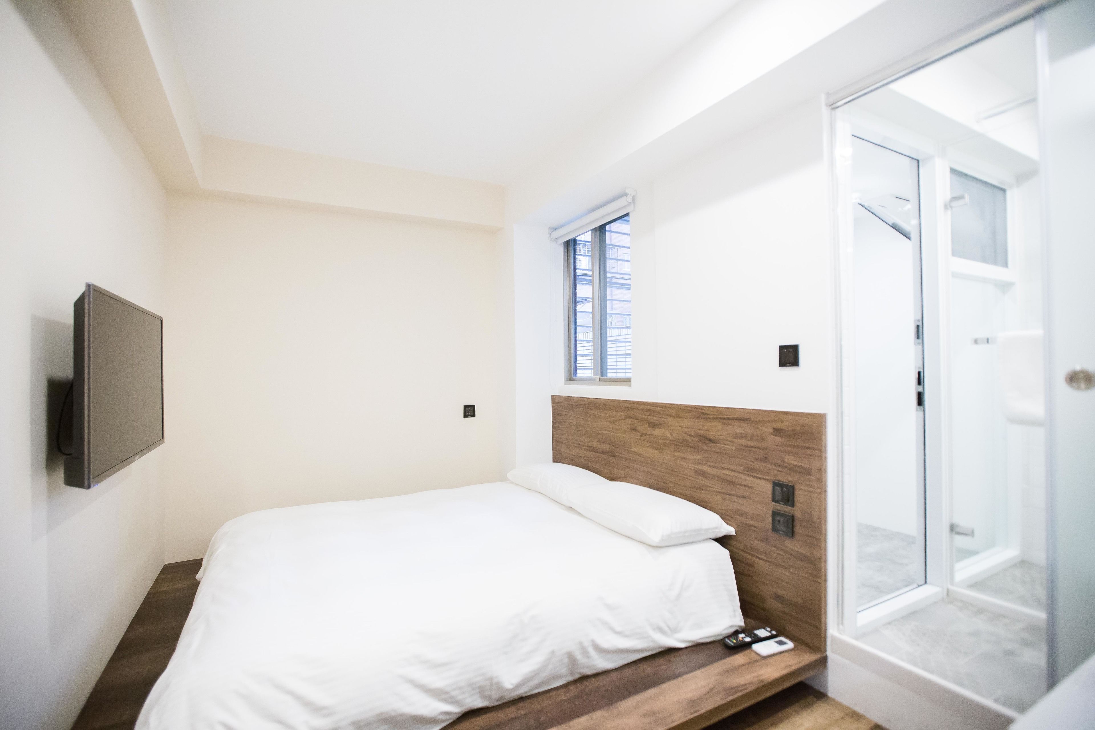 A modern bedroom with a wooden headboard, a bed with white bedding, a mounted TV, and a sliding glass door leading to a shower area.