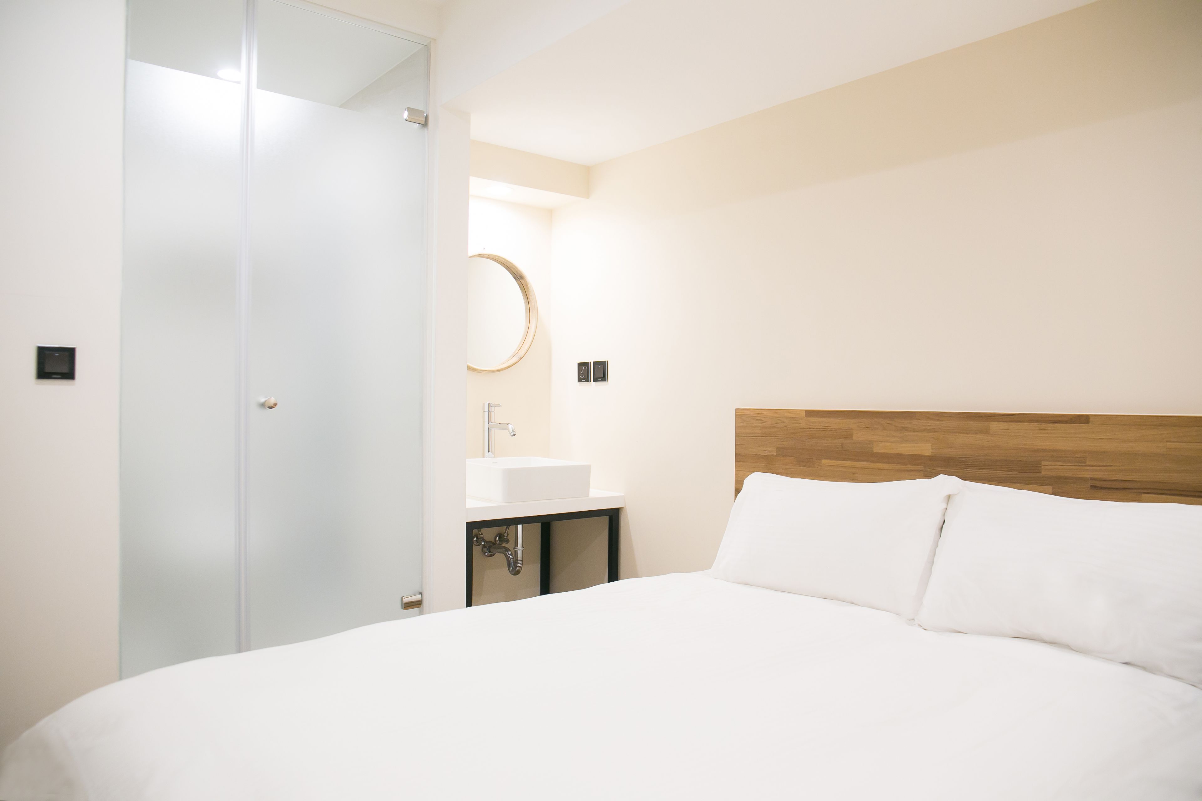 Habitación de hotel con cama, cabecero de madera, espejo redondo y mesita de noche con lavamanos.