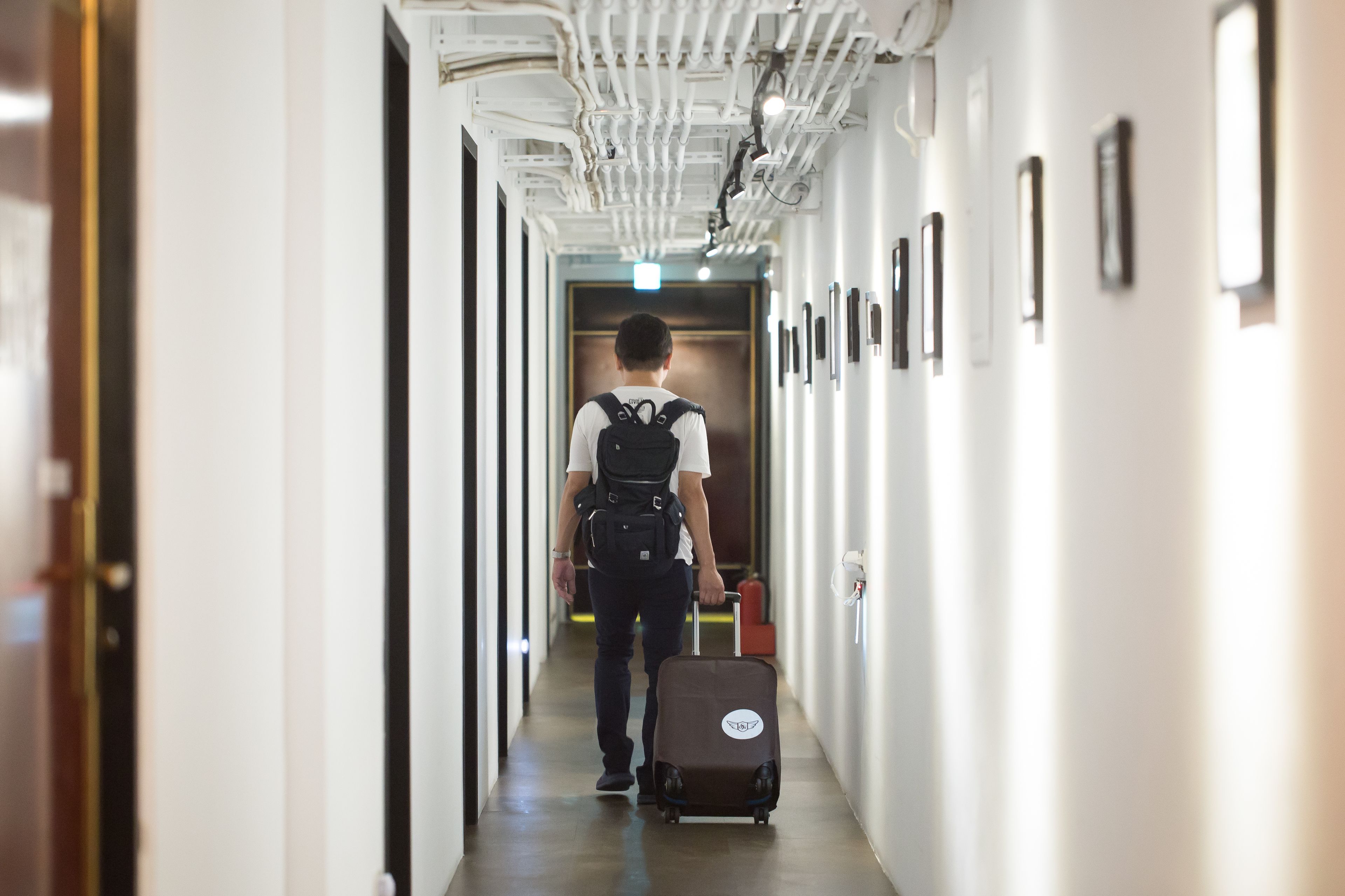 Corredor de hotel com uma pessoa caminhando em direção a uma porta, carregando uma mala.