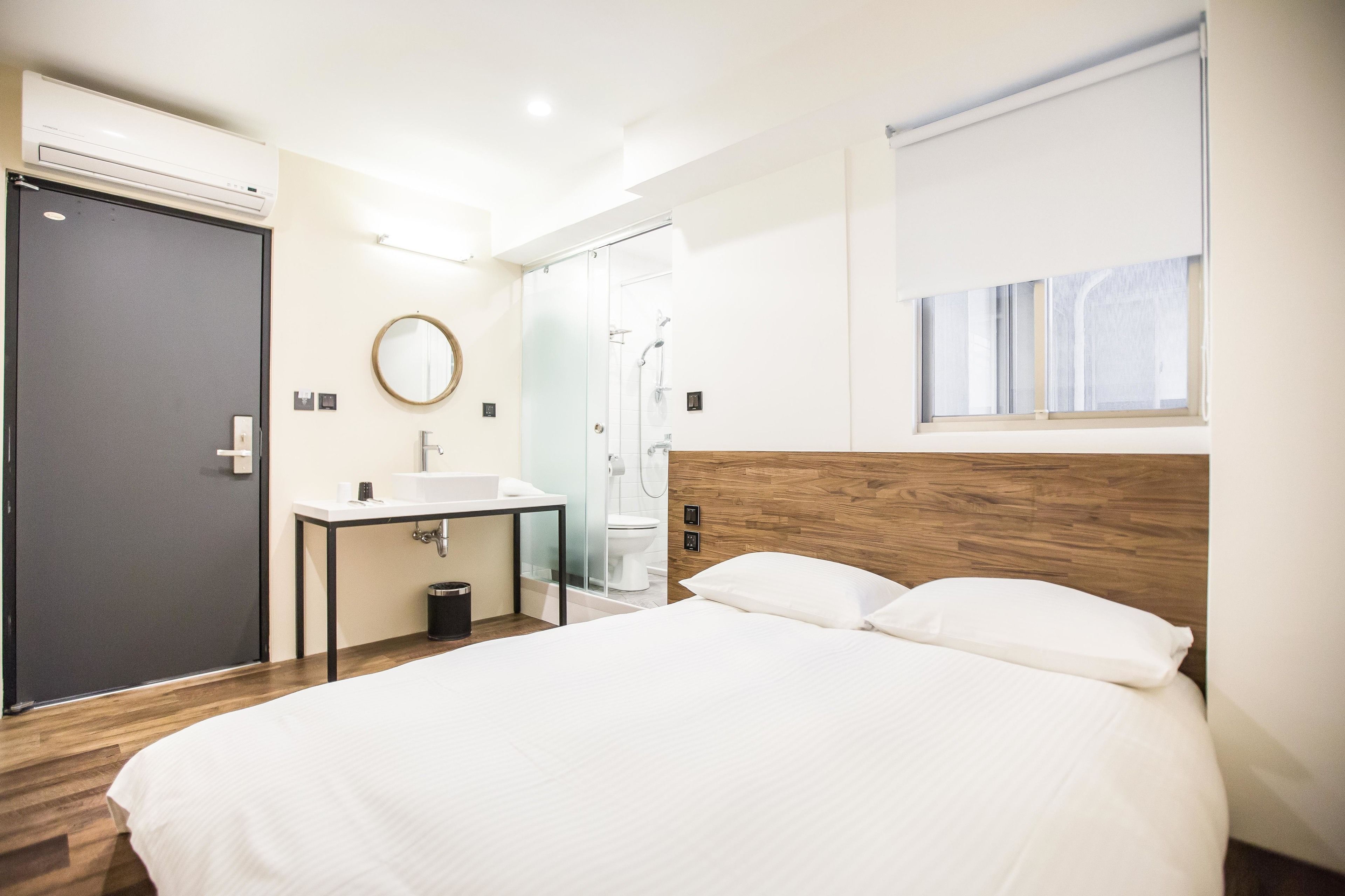 A modern hotel room with a large bed, a bedside table, a built-in sink, a mirror, and a shower area.