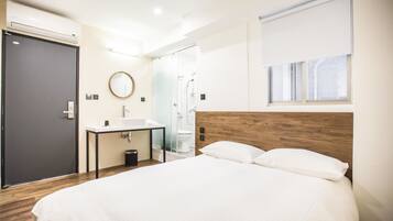 A modern hotel room with a large bed, a bedside table, a built-in sink, a mirror, and a shower area.