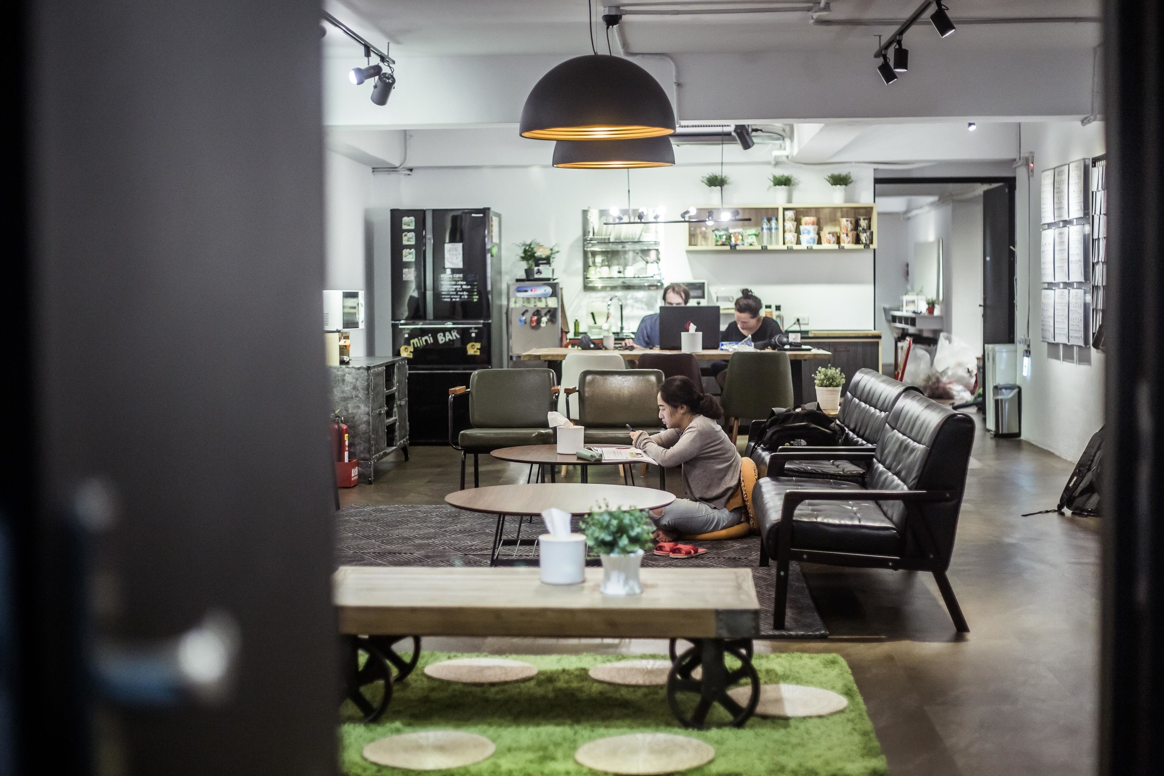 Un área de estar moderna con un sofá, una mesa de centro y una mesa de comedor. Se observa una cocina al fondo con un refrigerador y una persona trabajando en una computadora.