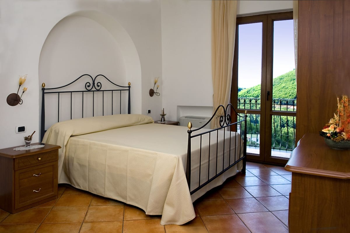 A bedroom with a bed, nightstand, dresser, and a view of greenery through a window.