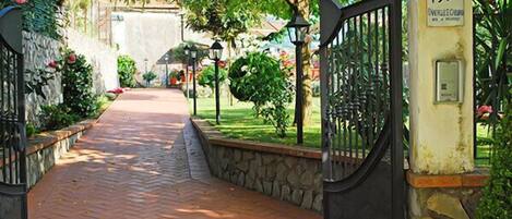 A paved pathway leading to a gated entrance with stone walls and greenery.