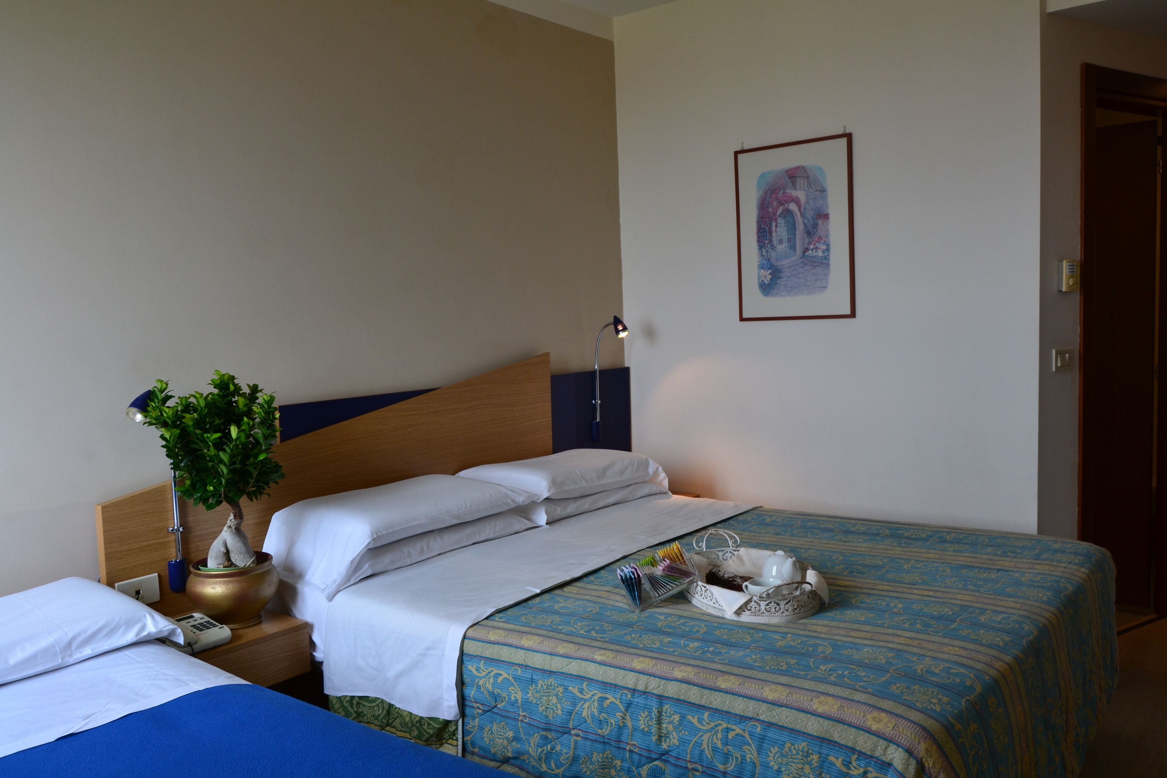 Une chambre d’hôtel avec deux lits, une table de chevet avec une lampe, une plante en pot et un tableau encadré au mur.