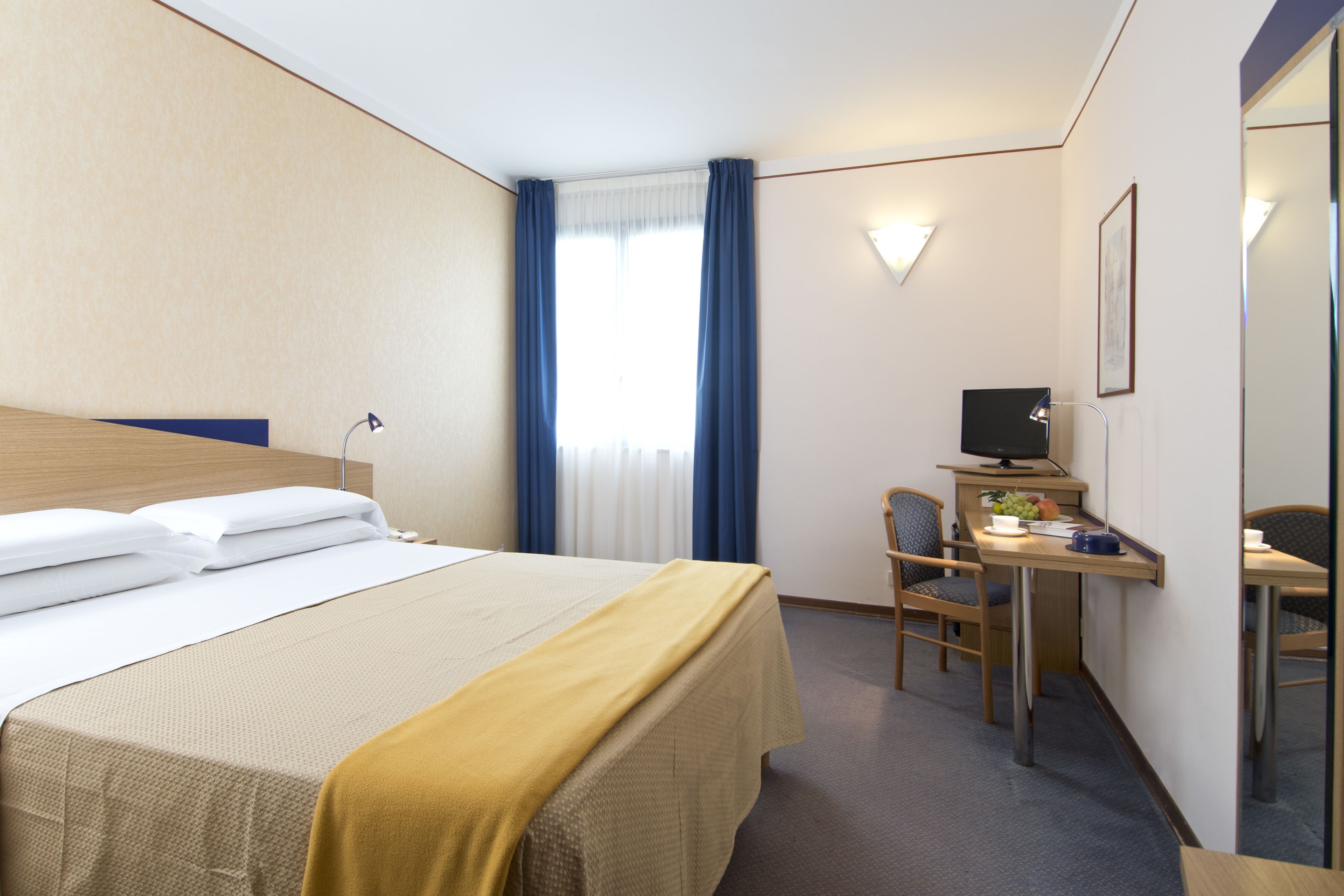 Una habitación de hotel con cama, escritorio, silla, televisor y ventana con cortinas.