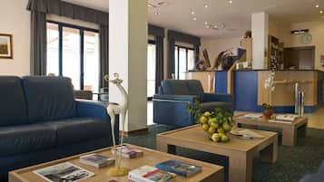 Un hall de hotel con un sofá azul, una mesa de centro de madera con libros y un jarrón, y una recepción con un reloj.
