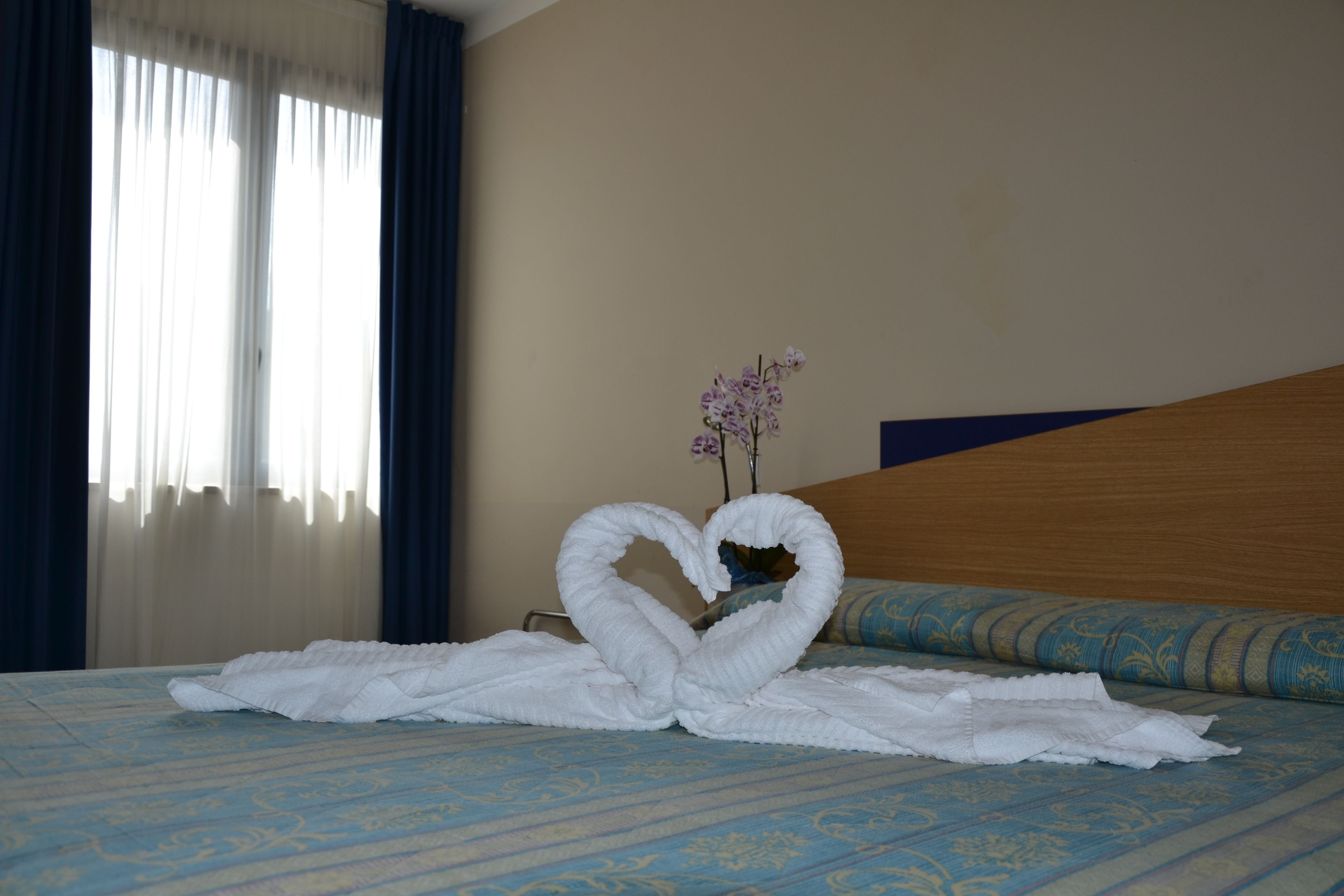 Une chambre d’hôtel avec un lit, une décoration en forme de cœur réalisée avec des serviettes, un vase rempli de fleurs et une fenêtre avec des rideaux.