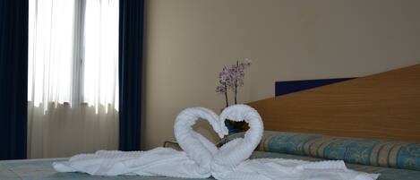 Une chambre d’hôtel avec un lit, une décoration en forme de cœur réalisée avec des serviettes, un vase rempli de fleurs et une fenêtre avec des rideaux.