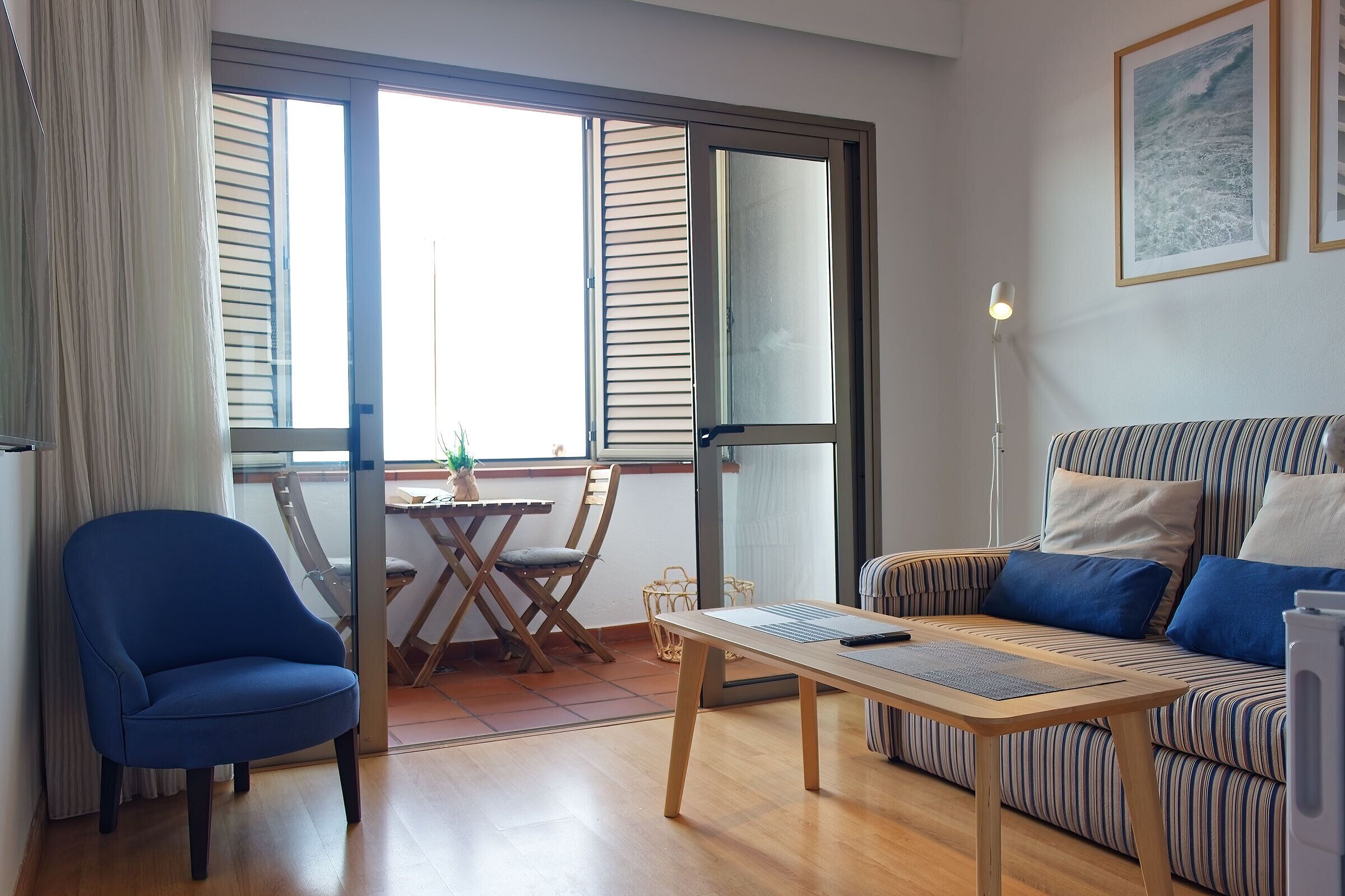 A modern living room with a blue armchair, a striped sofa, a wooden coffee table, and a large window with blinds.