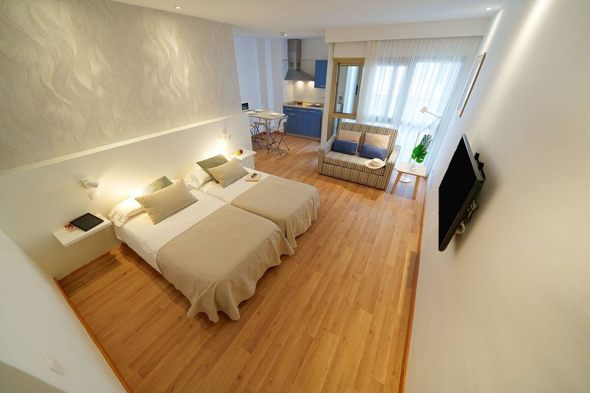 A modern hotel room with two beds, a desk, a chair, a TV, and a view of the kitchen and living area.