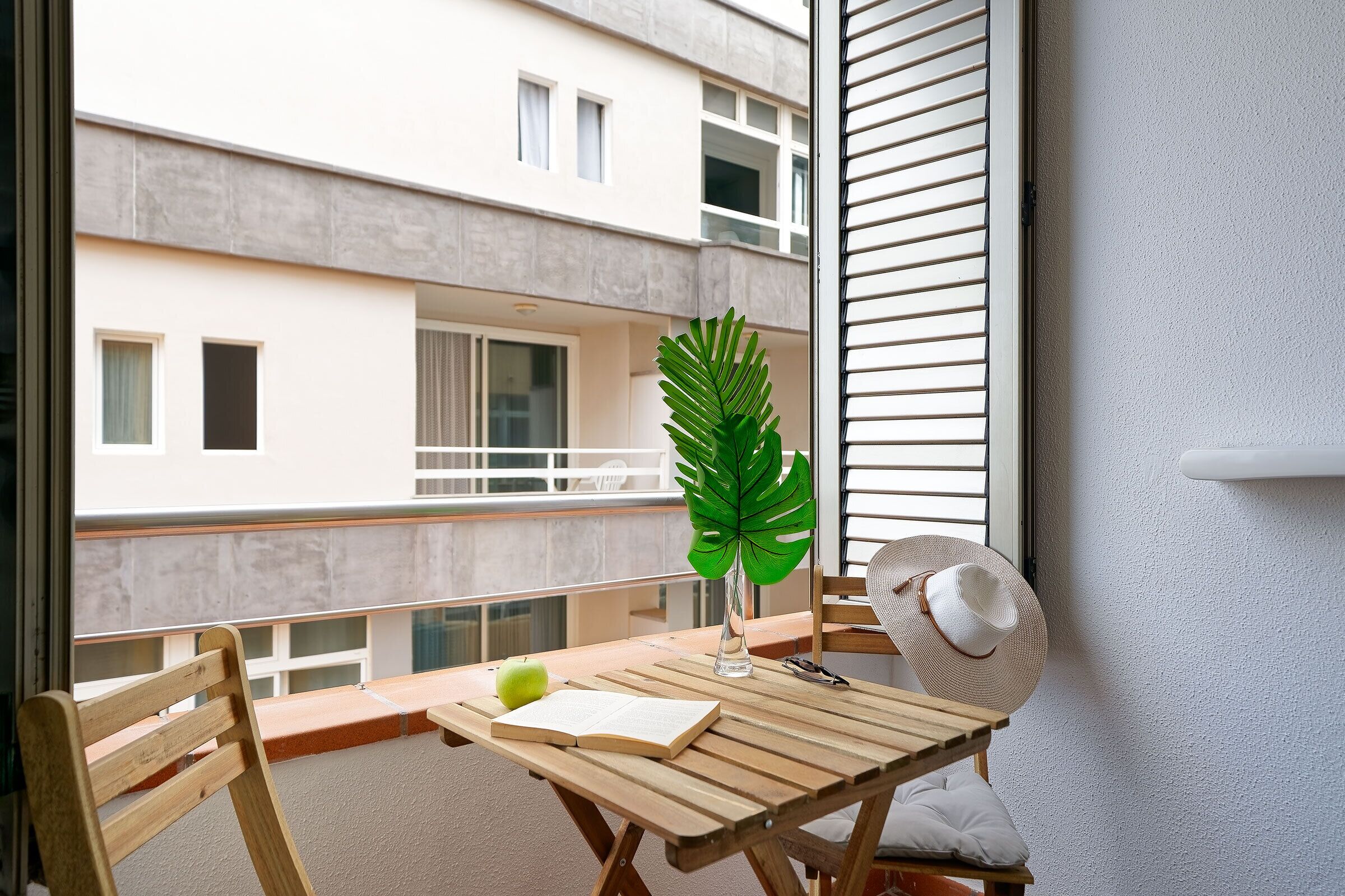 A balcony with a wooden table, a chair, a hat, a book, a plant, and a window overlooking a building.