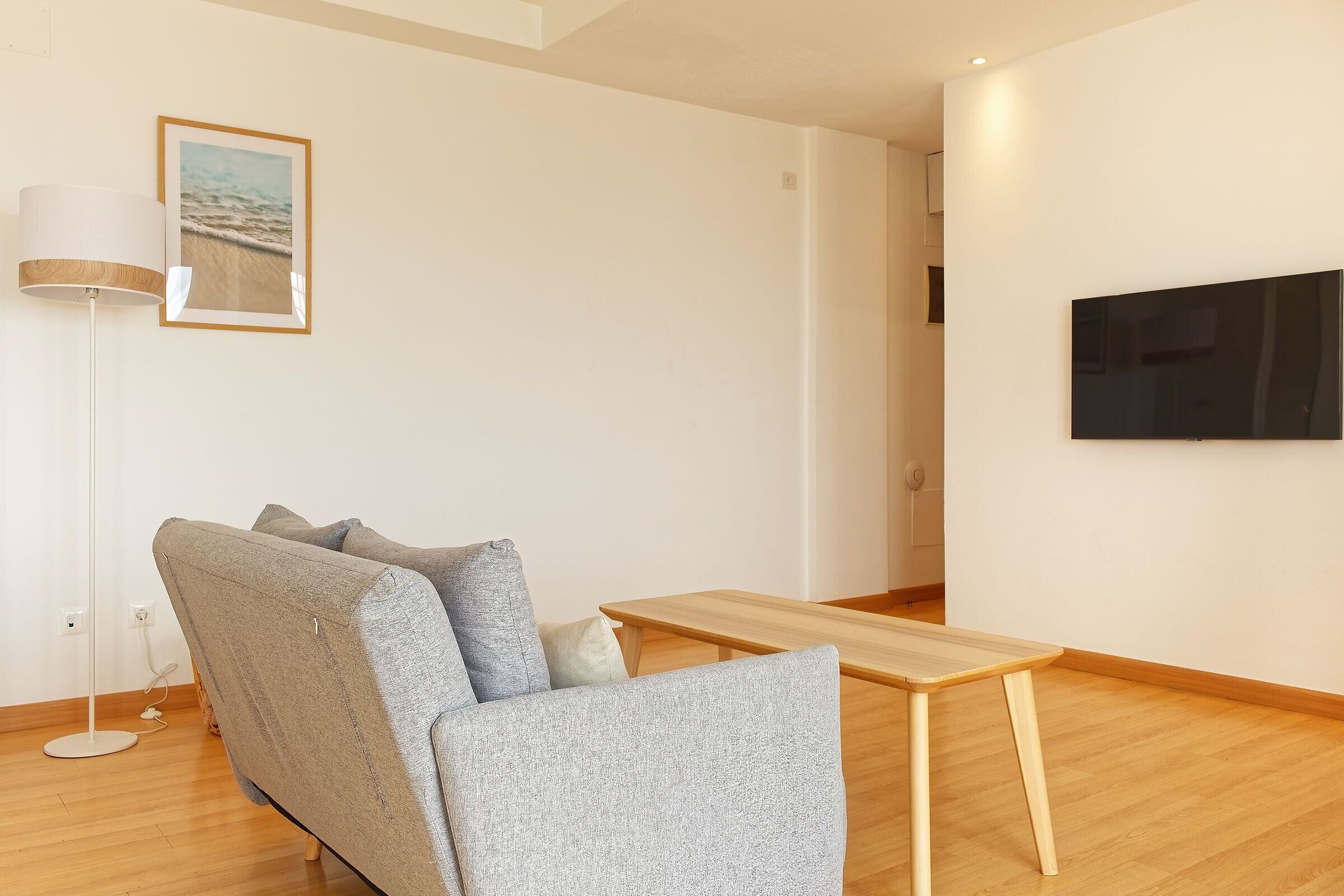 Uma sala de estar moderna com um sofá cinzento, uma mesa de centro de madeira, um abajur e uma televisão de tela plana presa à parede.