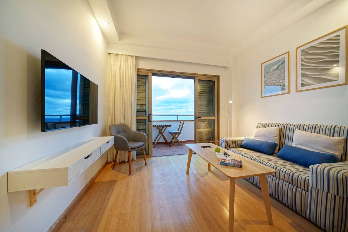 A modern living room with a sofa, a coffee table, a TV mounted on the wall, and a balcony with a view.
