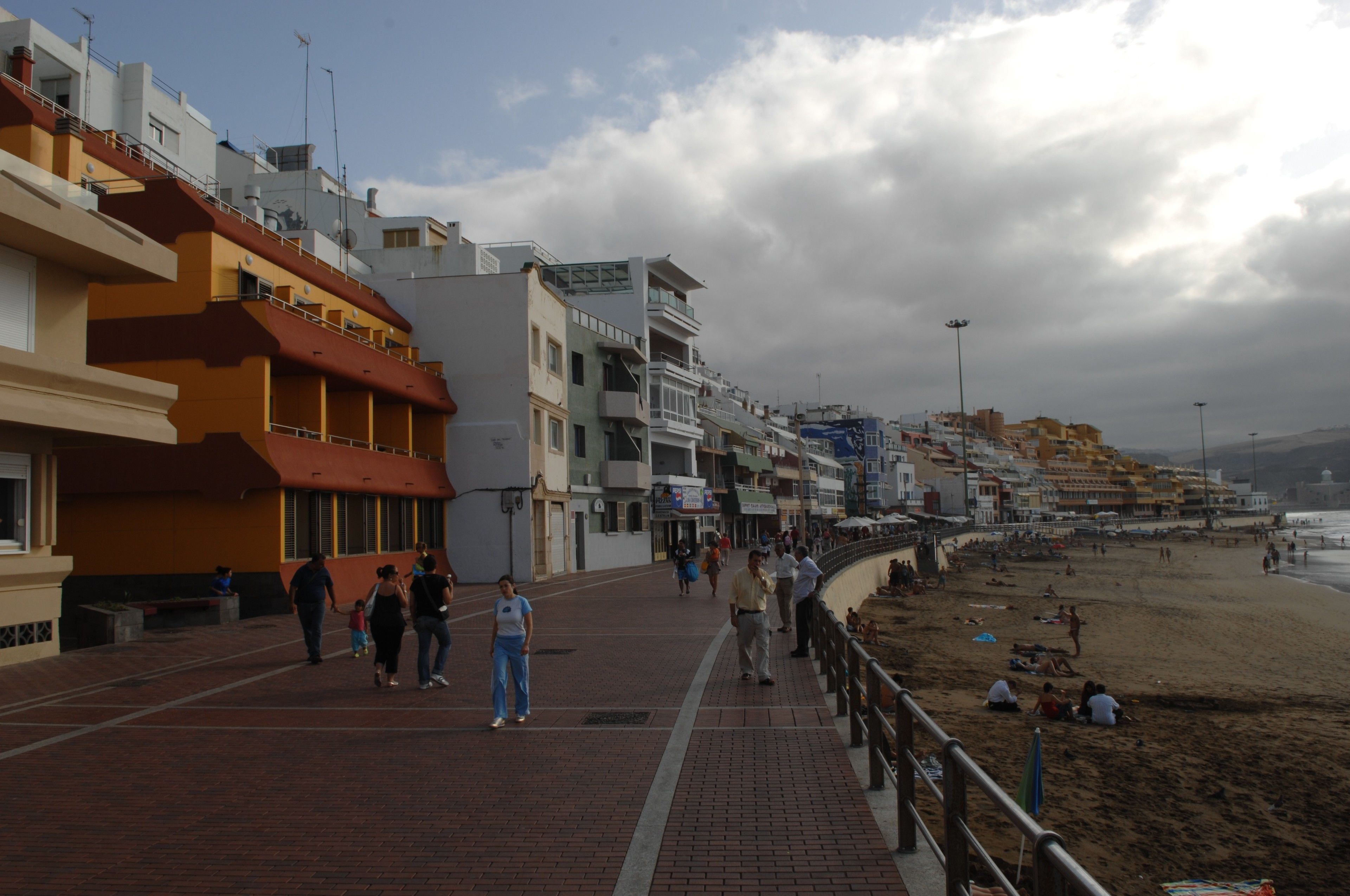A coastal promenade with buildings, a beach, and people walking.